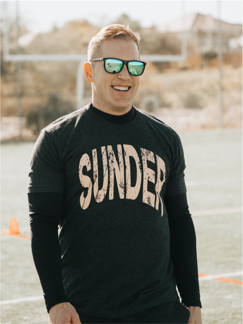 A man in sunglasses and a dark long sleeve shirt with the words 'Sunder' outside at a sporting event